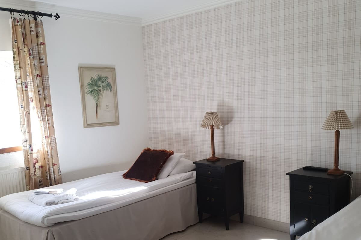 A clean, well-lit bedroom with two beds, a nightstand, a lamp, and a wall-mounted picture.