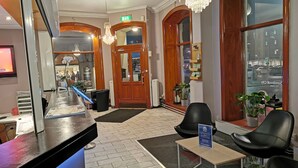 Un hall d'hôtel comprenant un comptoir d'accueil, des fauteuils en cuir noir, un lustre et une vue sur la rue grâce à de grandes fenêtres.