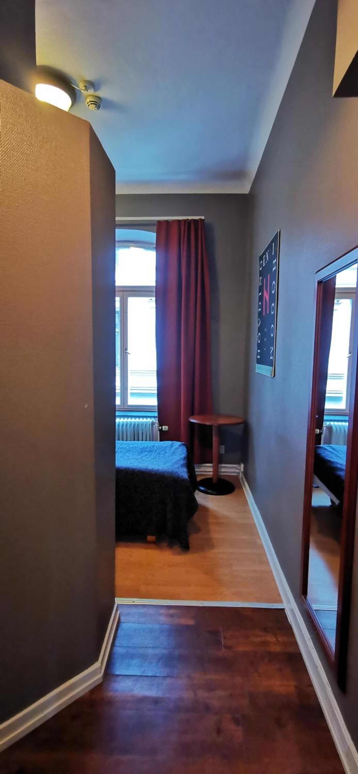 En smal gang med en seng for enden, et lille bord og et spejl på højre væg.