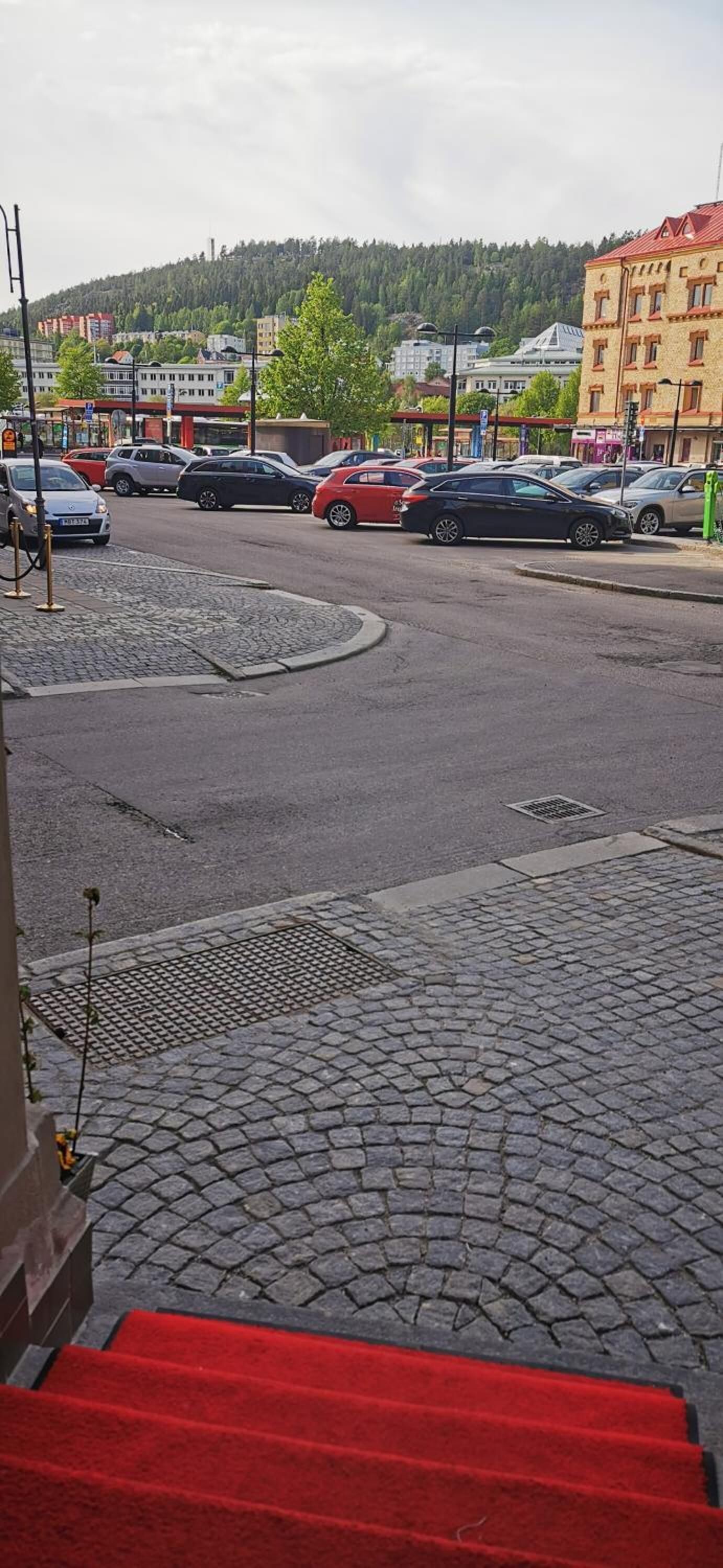 A street view with a red carpet leading to a building entrance, parked cars, and a hilly background.