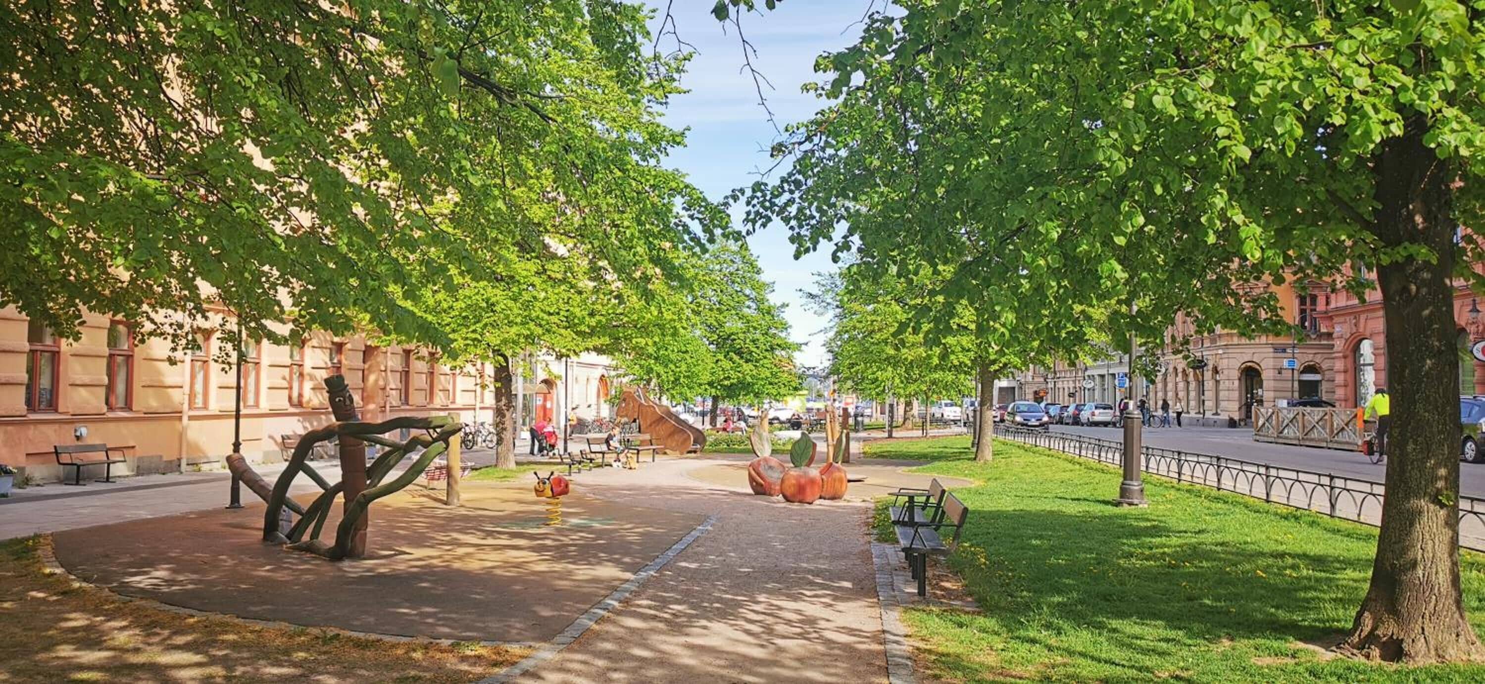 A tree-lined pedestrian path with benches, a sculpture, and a playground.