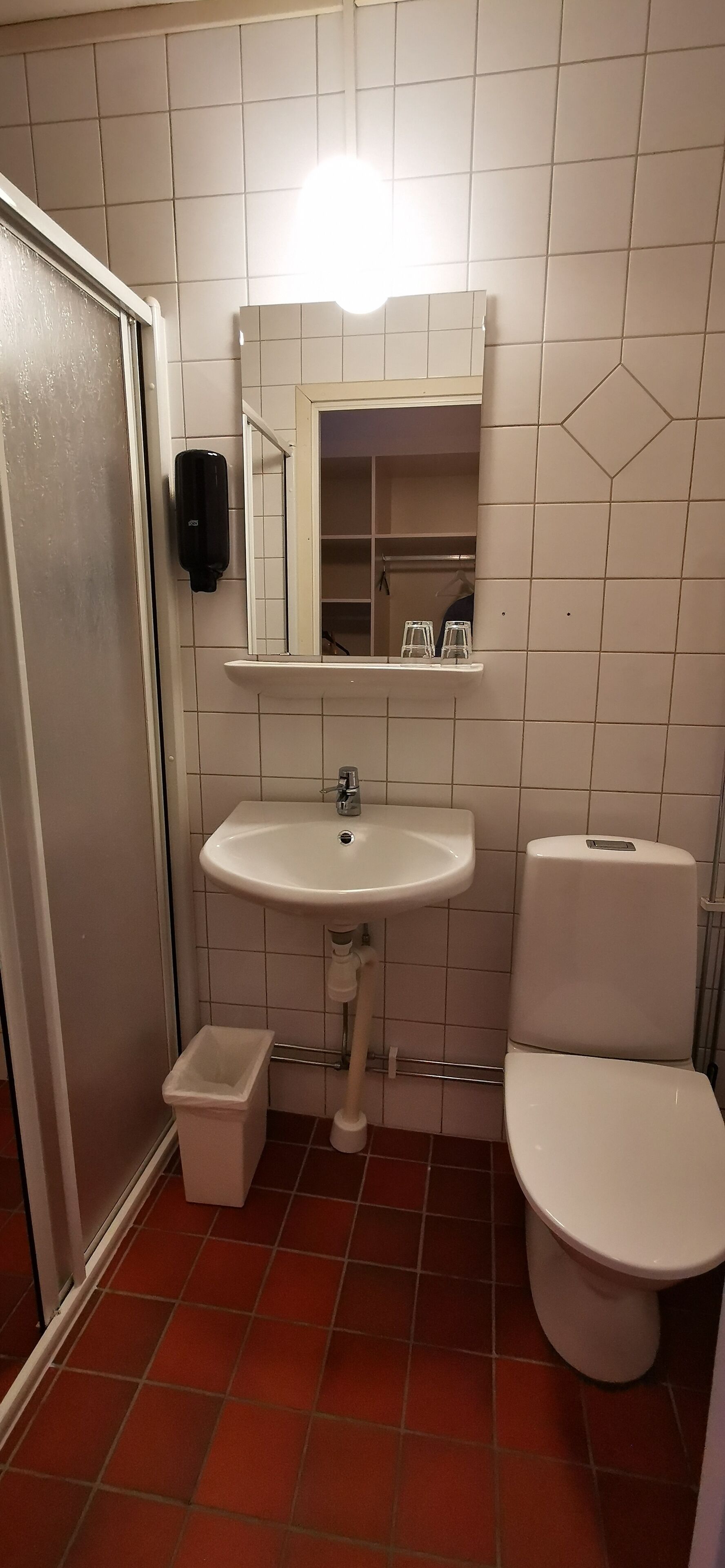 Un baño con lavamanos blanco, espejo e inodoro. Hay un cubo de basura al lado del lavamanos y un estante encima del espejo.