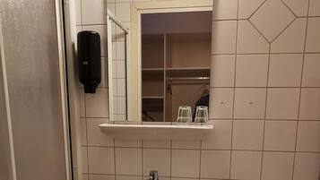 Un baño con lavamanos blanco, espejo e inodoro. Hay un cubo de basura al lado del lavamanos y un estante encima del espejo.