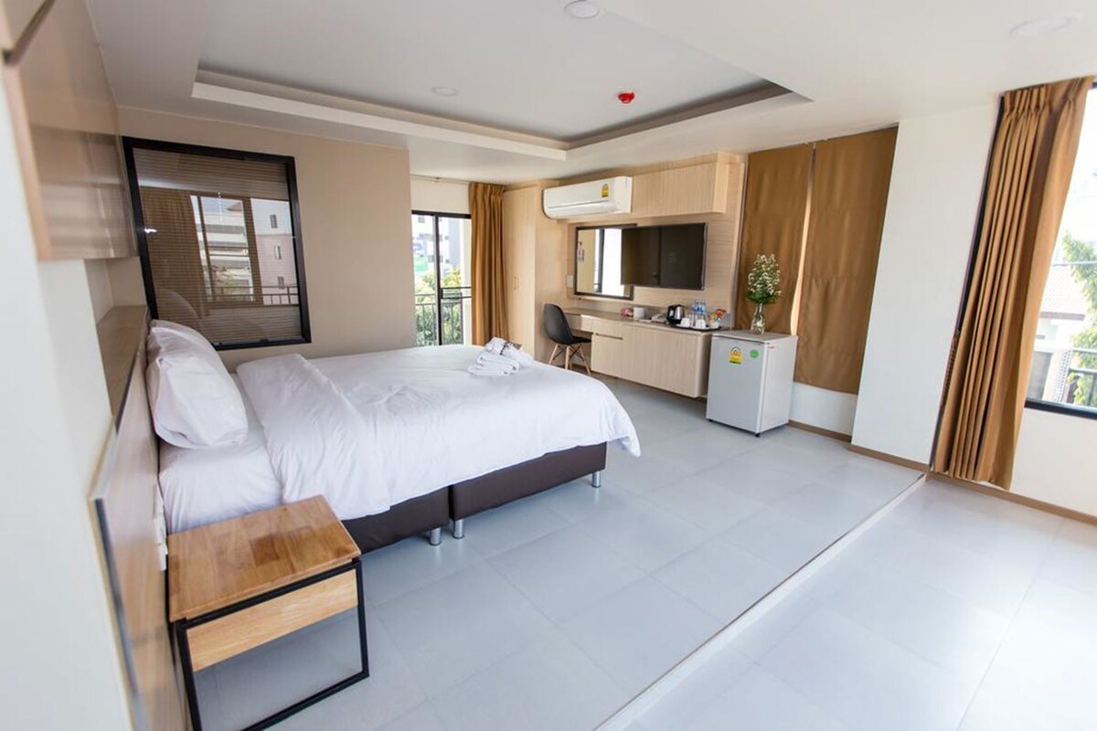 A modern hotel room with a large bed, a desk, a TV, and a kitchenette.
