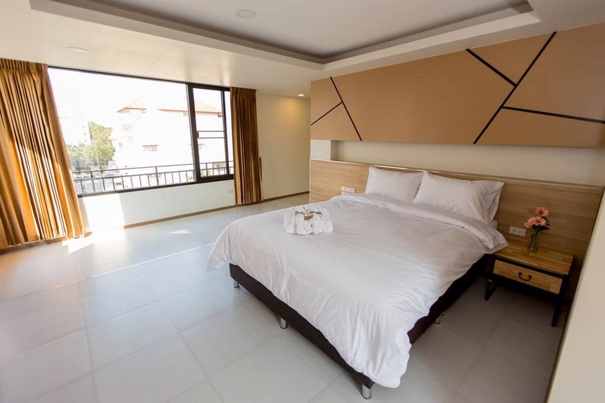 Habitación de hotel moderna con una cama grande, una mesita de noche con un jarrón de flores y vistas al exterior a través de amplias ventanas.