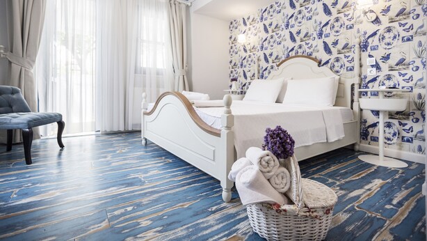 A bedroom with a white bed, a blue and white patterned wall, a white nightstand, a blue and white striped floor, and a grey armchair. - Sardunya Otel (Bozcaada)