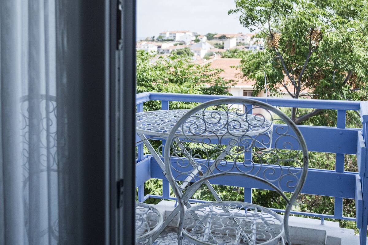 A balcony with a metal table and chairs, a blue railing, and a view of a cityscape with buildings and trees.