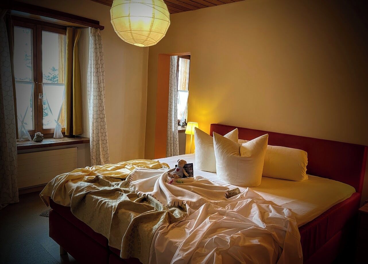 Una cama bien tendida con almohadas y una colcha, una lámpara de noche y una ventana con cortinas.