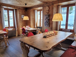 A wooden cabin interior with a large dining table, multiple chairs, and a hanging lamp.