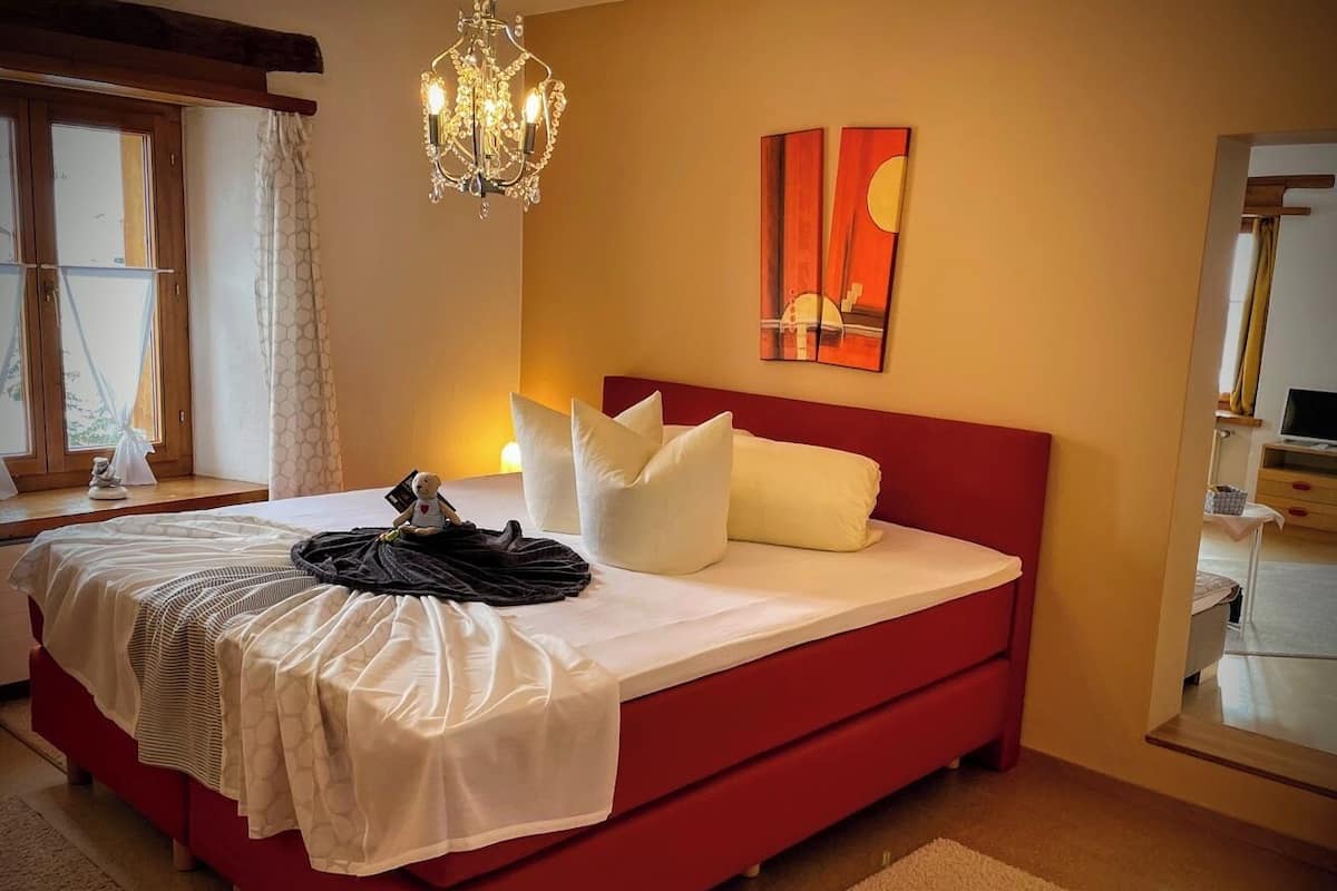 A bedroom with a red bed, white bedding, a chandelier, and a painting on the wall.