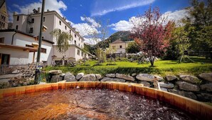A stone-lined outdoor pool with a wooden deck, surrounded by a stone wall and a grassy area with trees and buildings in the background. - Villa Stelvio Bed & Breakfast (Val Muestair)