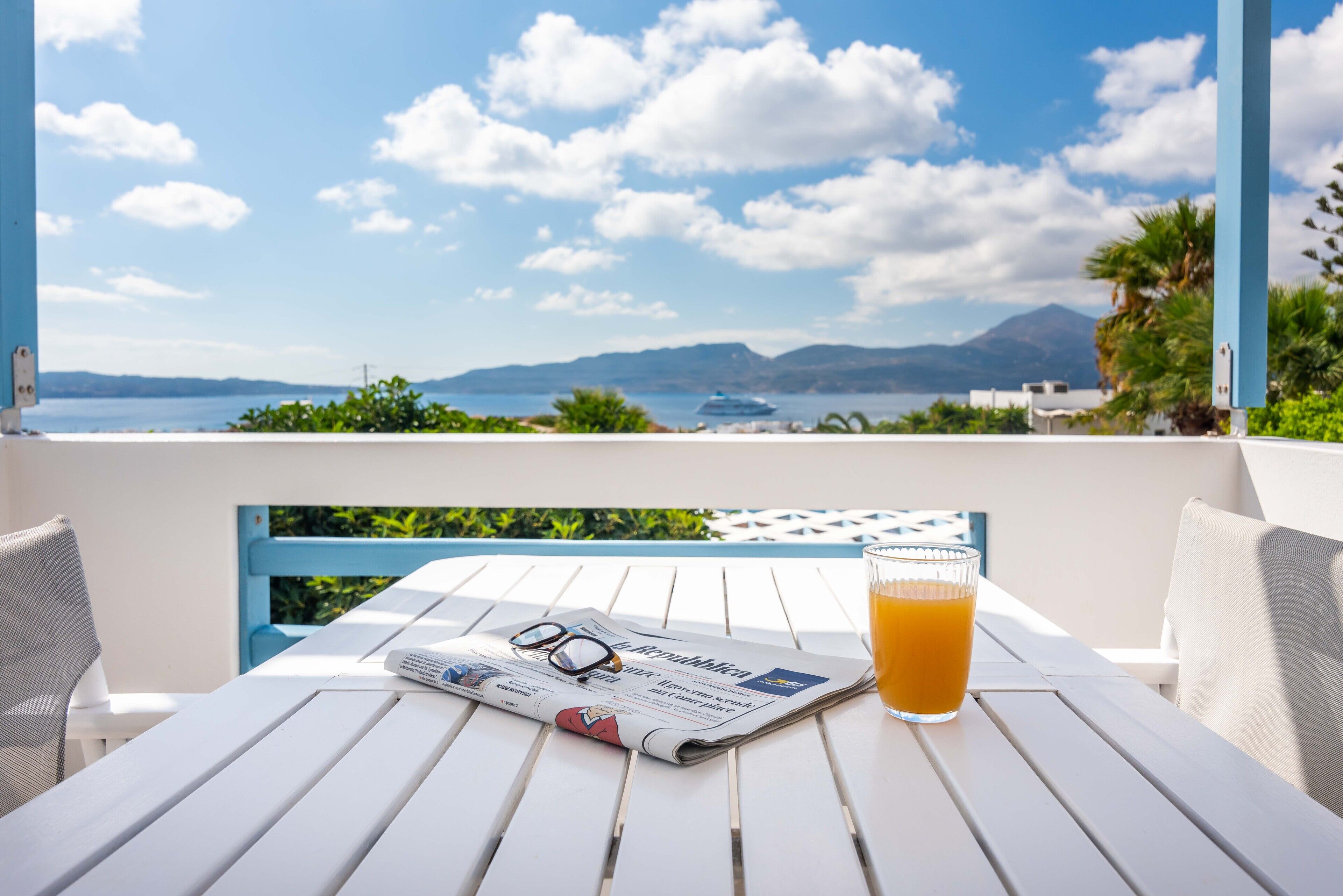 A white table with a newspaper, glasses, and a glass of orange juice, overlooking a scenic view of the sea and mountains.