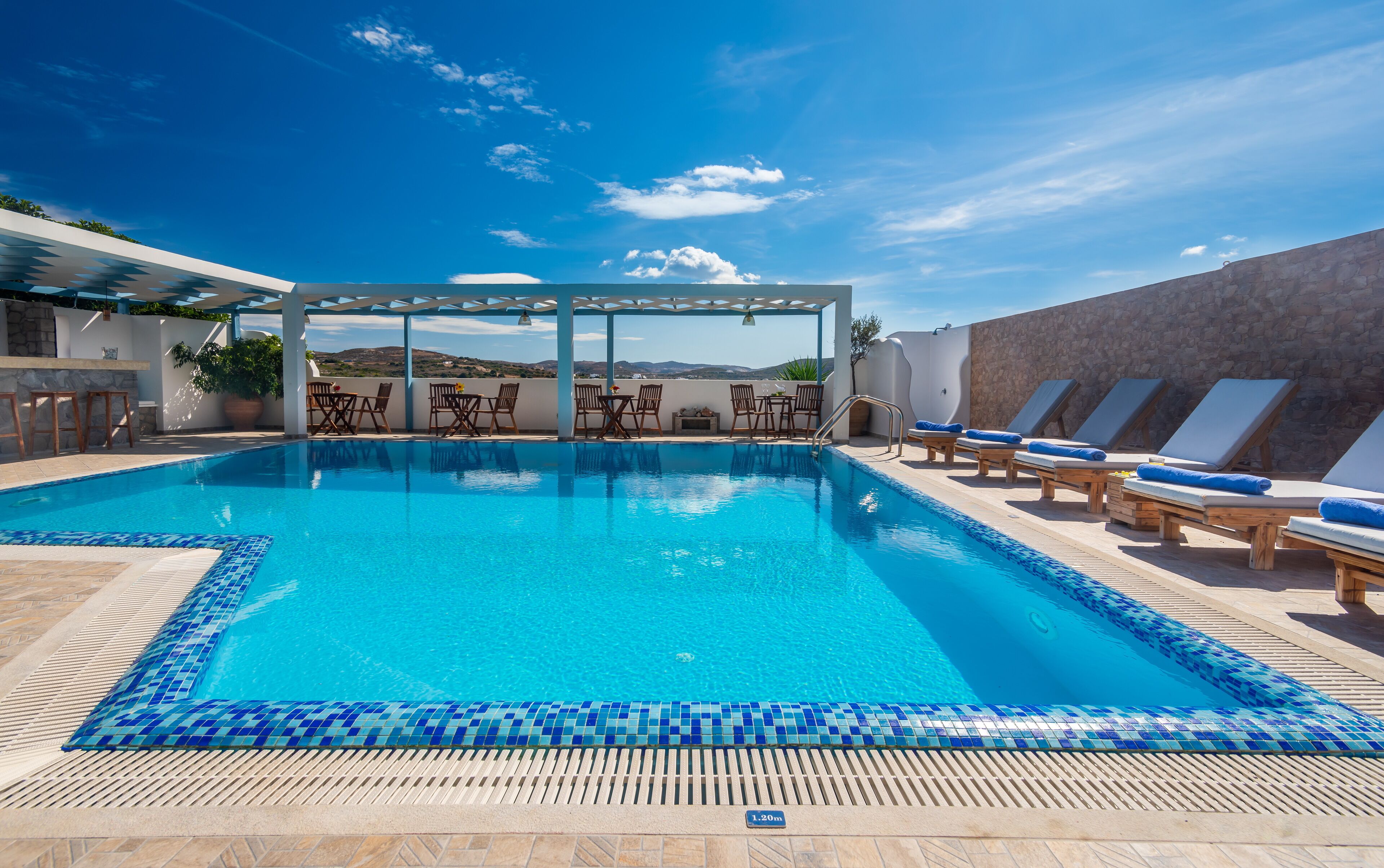 A pool area with a clear view of the sky, a pergola with chairs, and a stone wall.