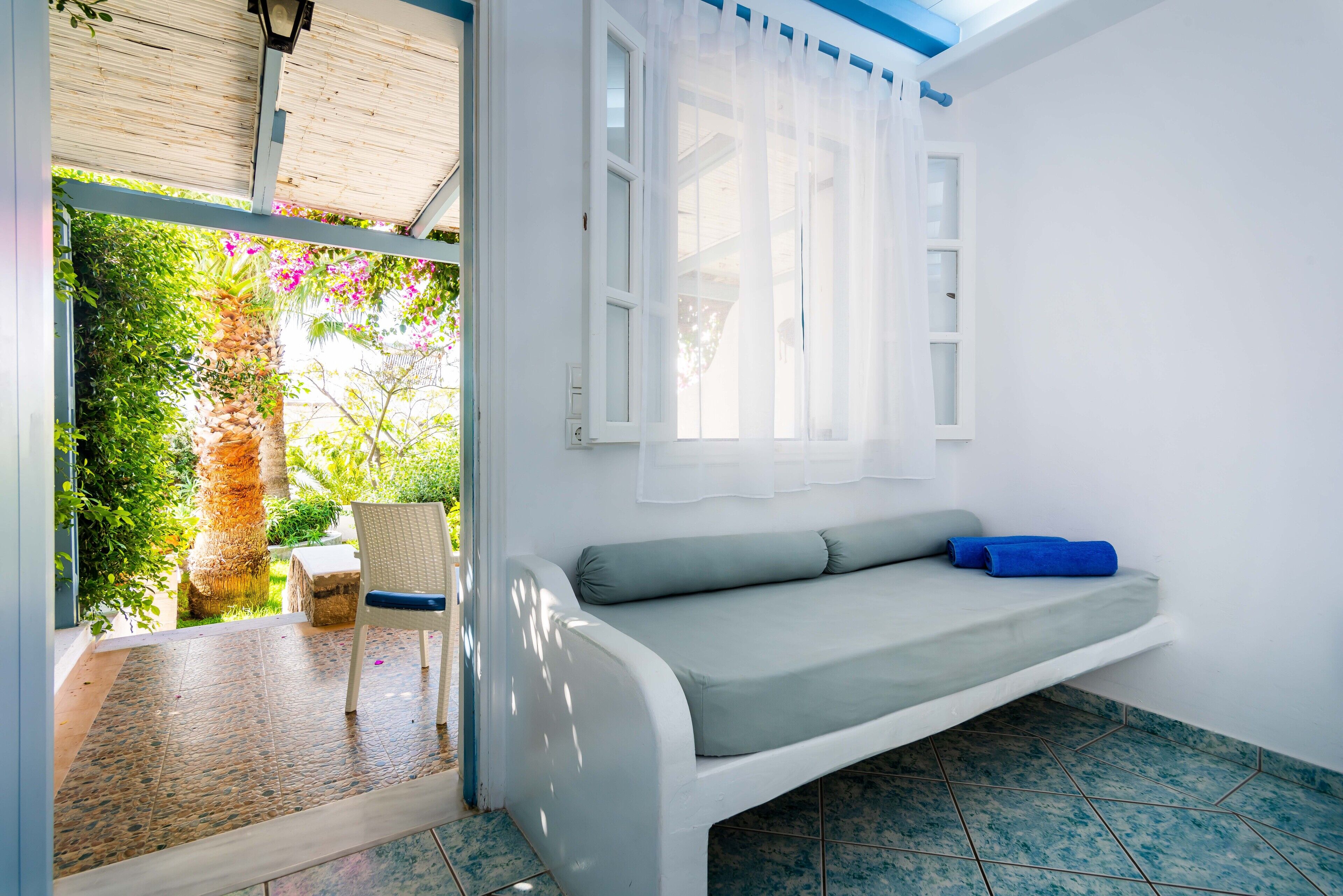 A room with a white sofa, a chair, a window with white curtains, and a view of greenery outside.
