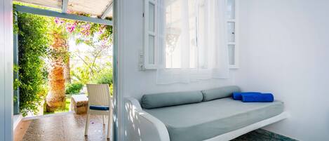 A room with a white sofa, a chair, a window with white curtains, and a view of greenery outside.
