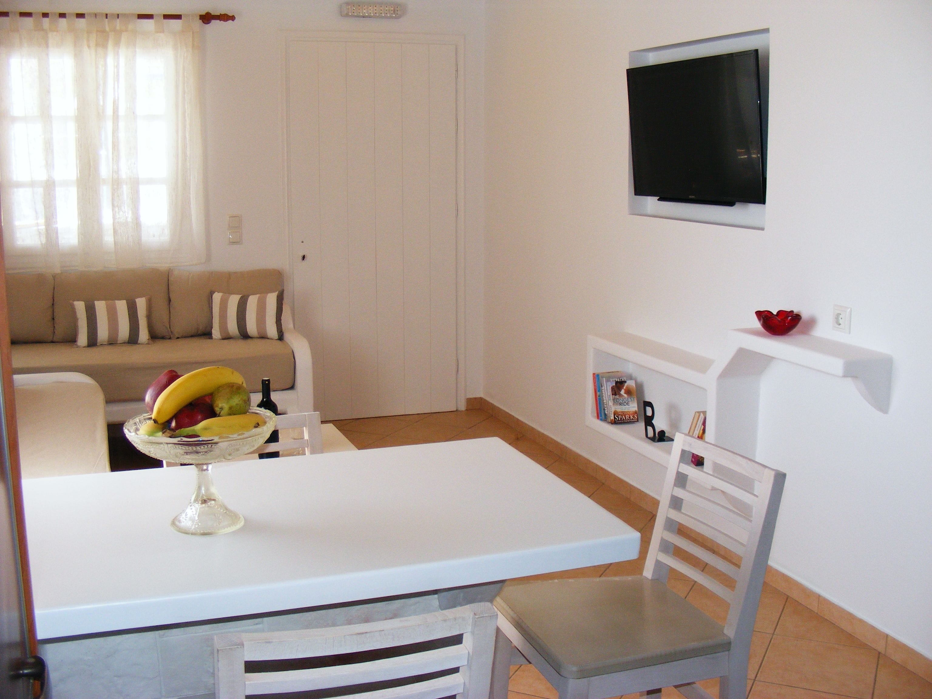 A living room with a white sofa, a dining table with a chair, a TV mounted on the wall, and a shelf with books and a bowl.