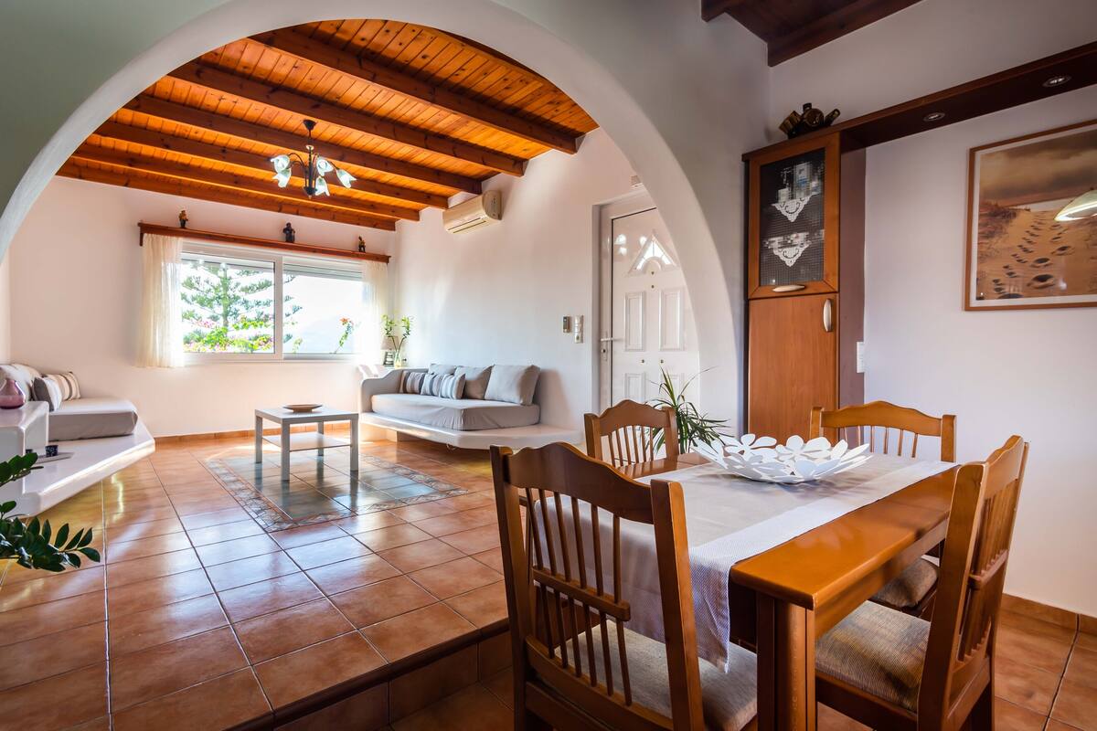 A dining area with a wooden table and chairs, a white sofa, and a window with a view of trees.