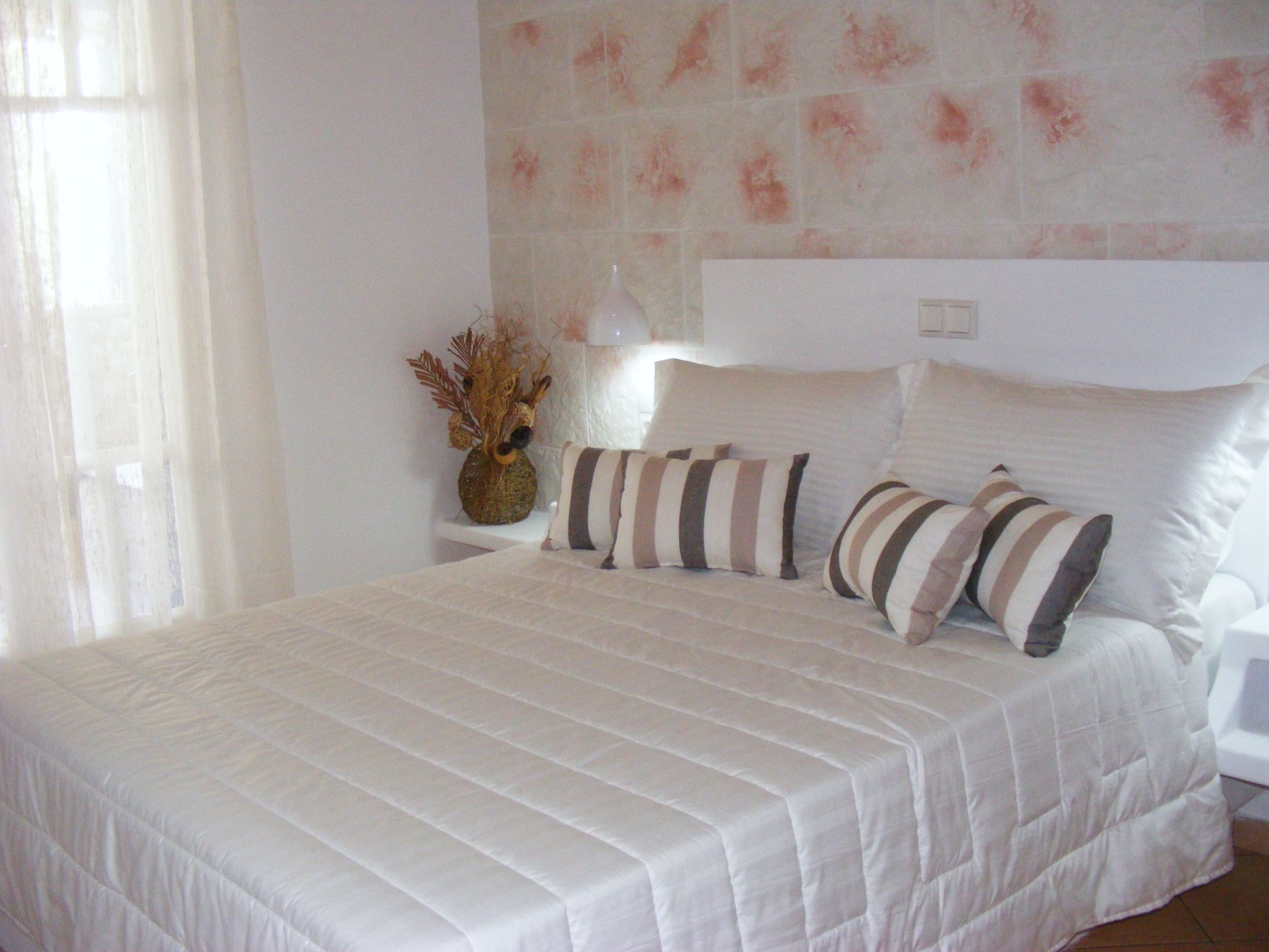 A neatly made bed with a striped bedspread and two pillows, a vase with dried plants on a bedside table, and a wall with a pinkish pattern.