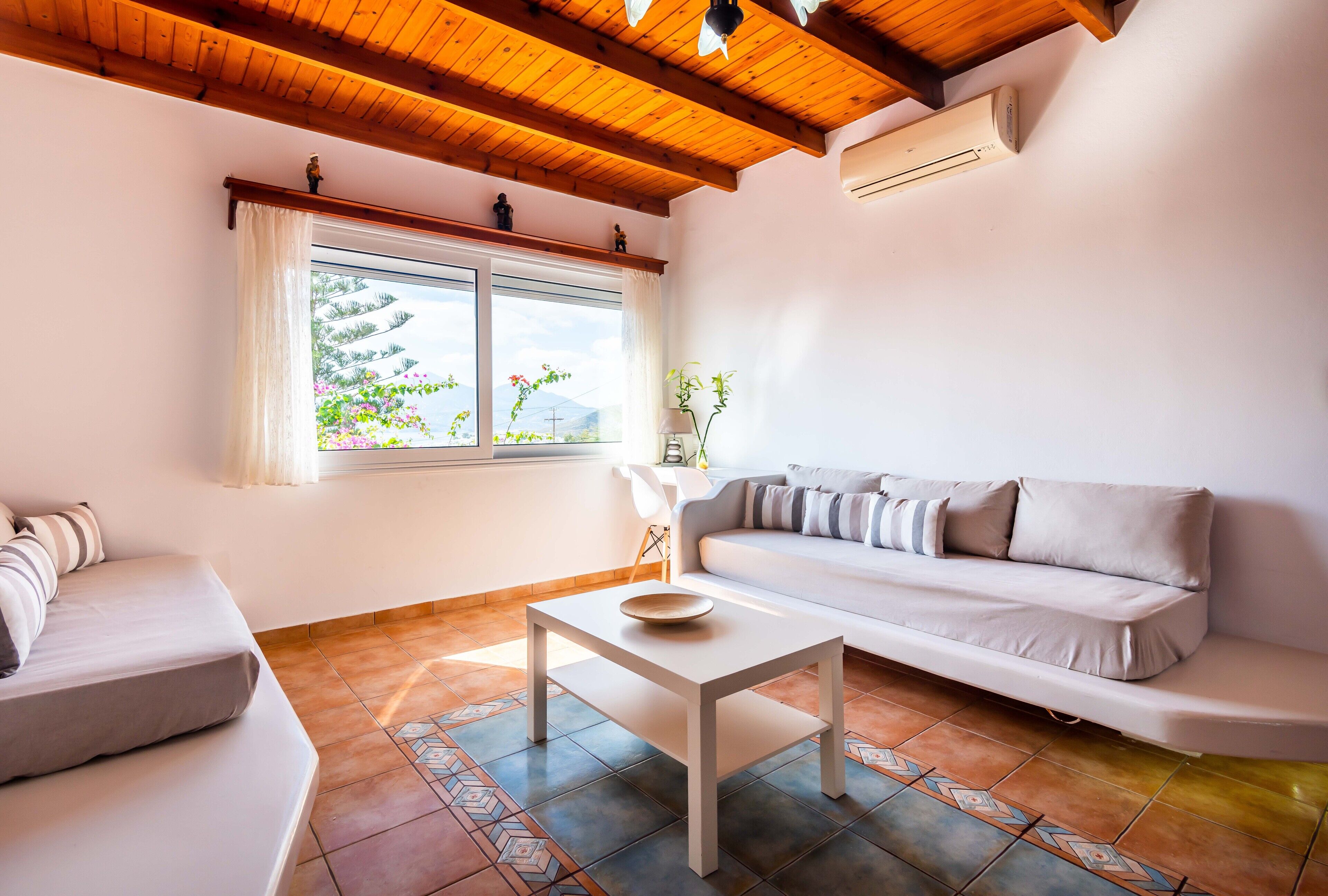 Ein modernes Wohnzimmer mit einem weißen Sofa, einem kleinen weißen Tisch und einem Fenster mit Blick auf Grünflächen und Berge.