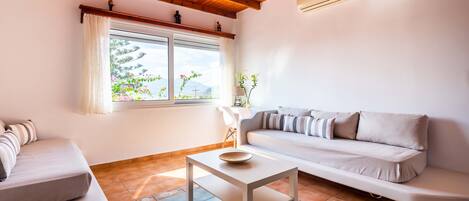 Ein modernes Wohnzimmer mit einem weißen Sofa, einem kleinen weißen Tisch und einem Fenster mit Blick auf Grünflächen und Berge.