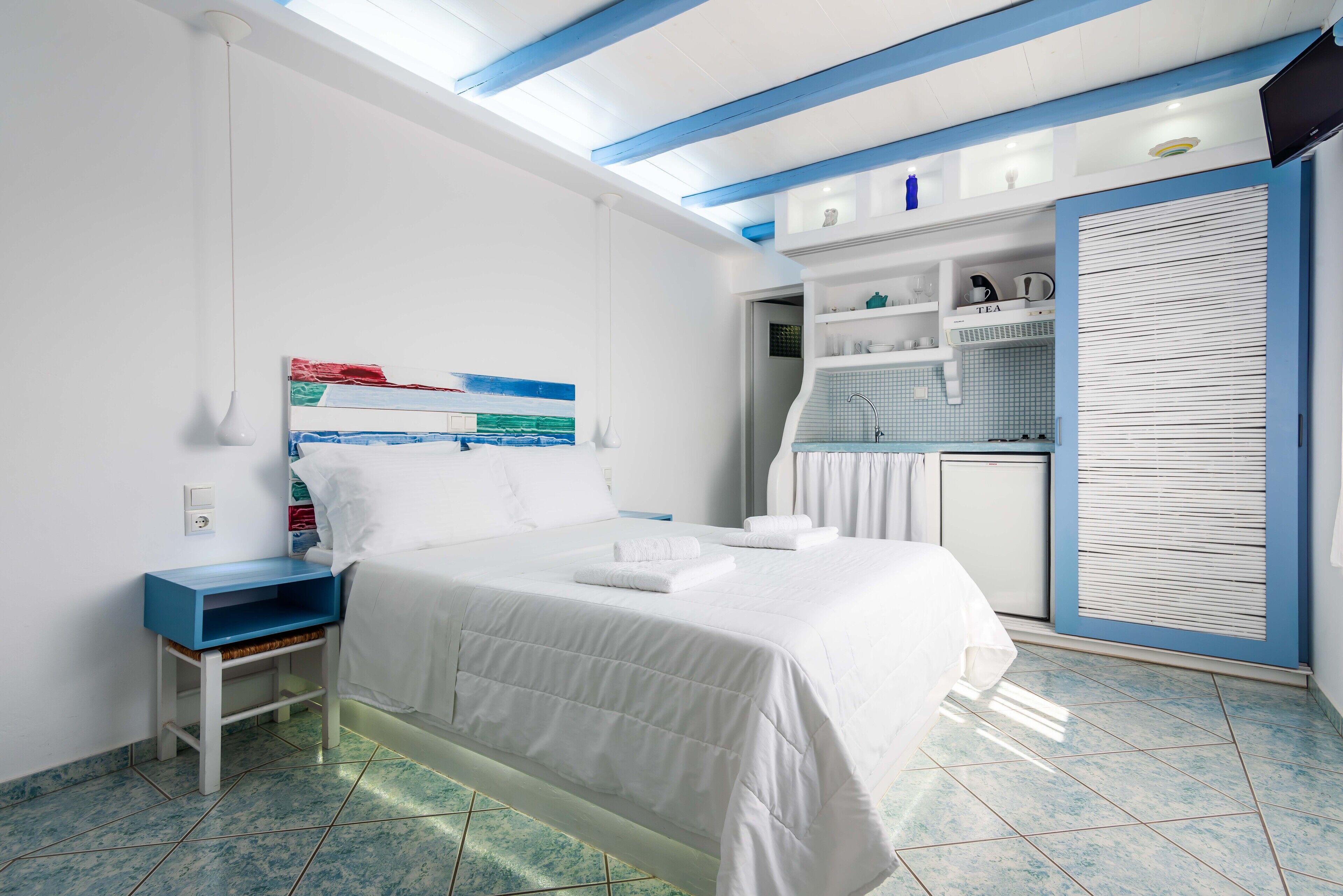 A bedroom with a bed, a nightstand, a shelf with toiletries, a window with blinds, and a kitchenette area.