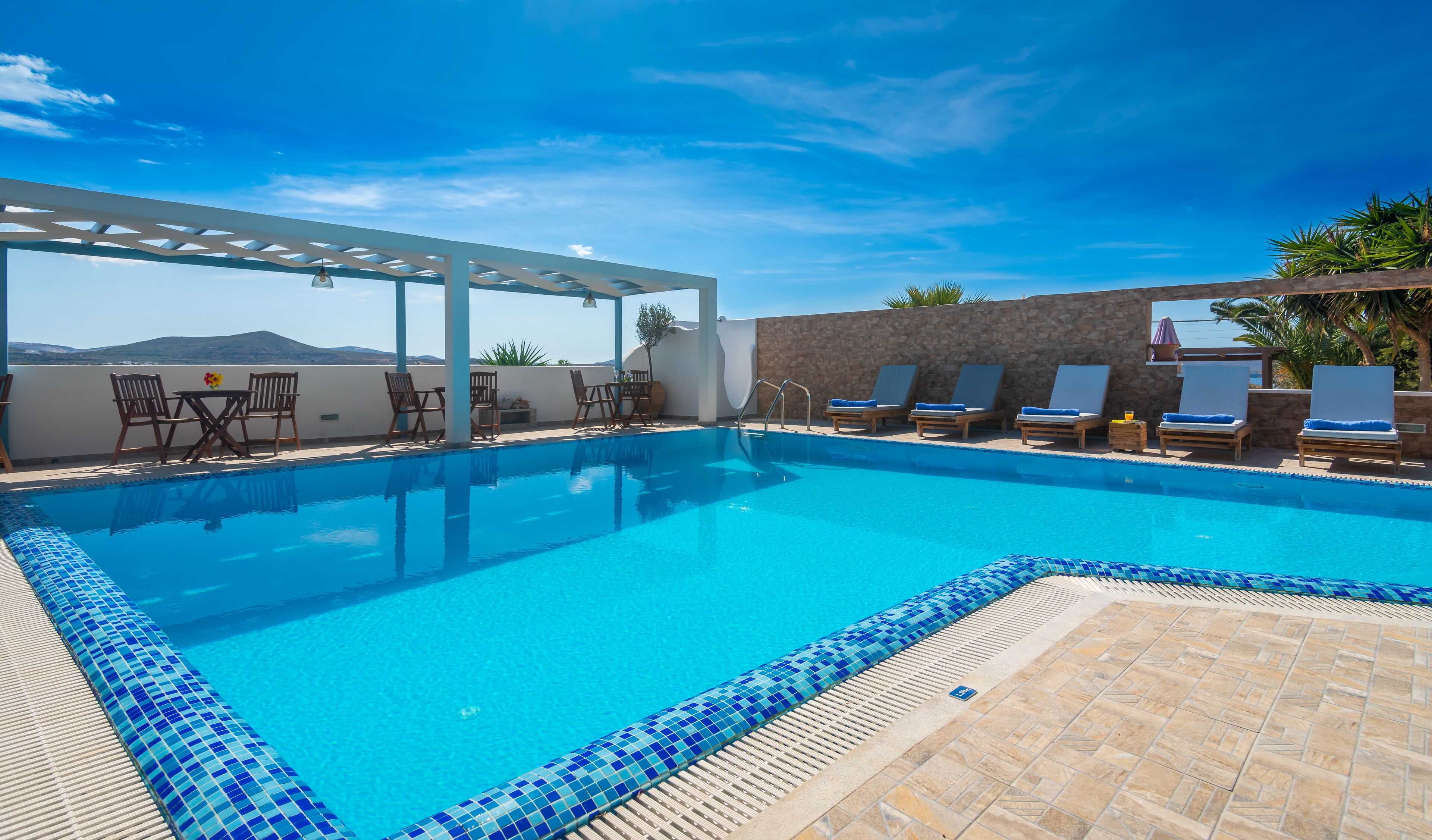 A clear blue pool with lounge chairs and a table, surrounded by stone walls and a pergola.