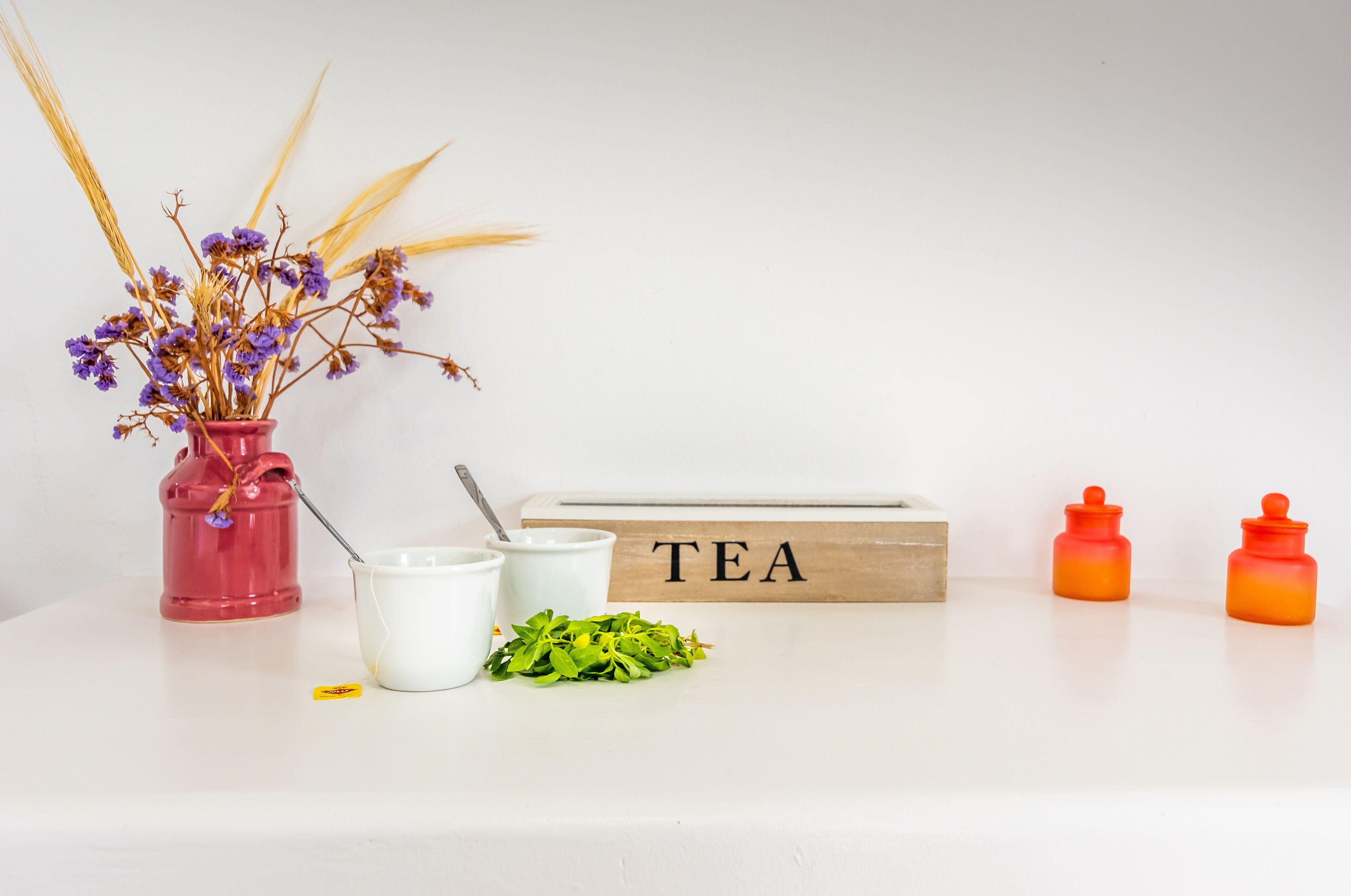 A white table with a red vase of dried flowers, a white cup, a wooden block with "TEA" written on it, and two orange containers.
