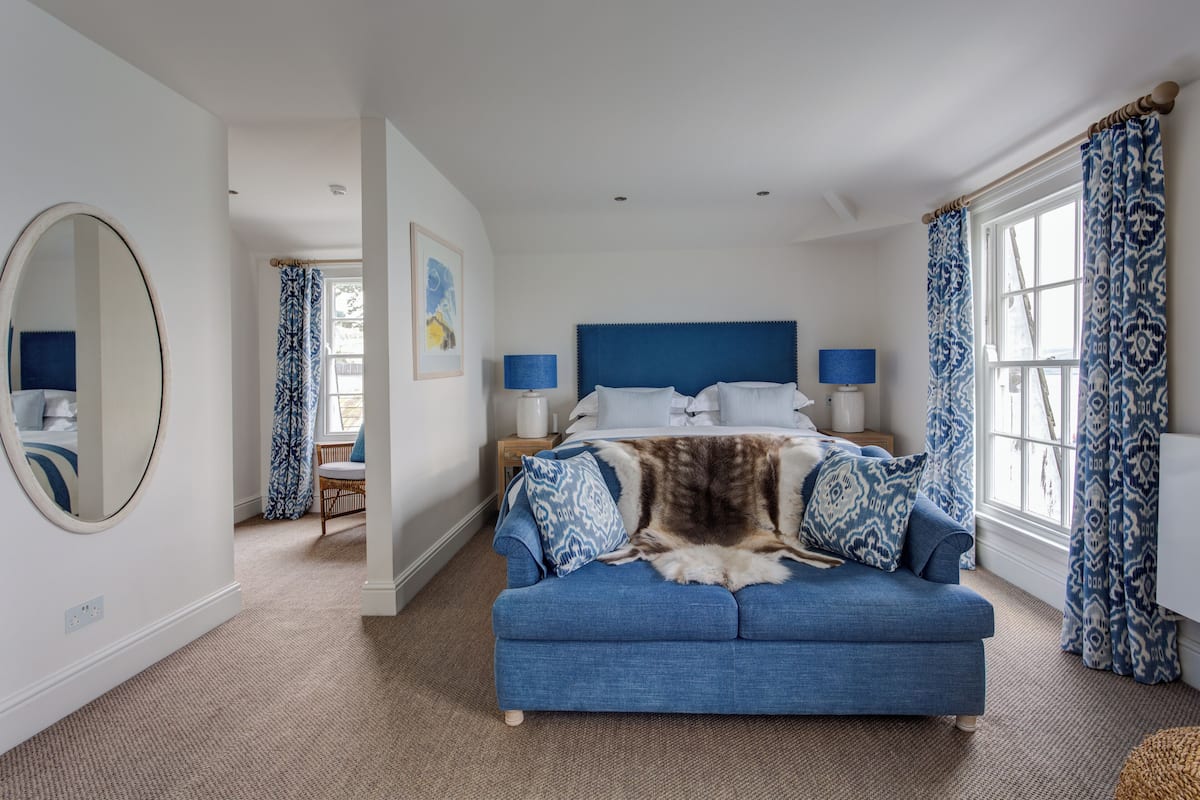 A bedroom with a large bed, a blue sofa, a round mirror, and a window with patterned curtains.
