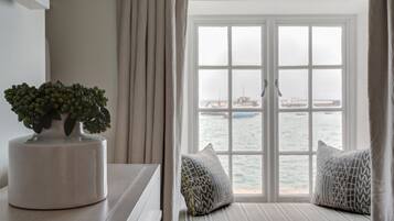 Quarto com vista para o mar através de uma grande janela, uma planta em vaso sobre uma mesa e um banco acolchoado com almofadas estampadas.