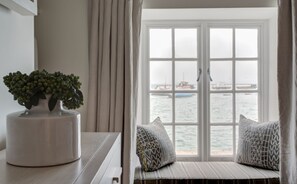 A room with a view of the sea through a large window, a potted plant on a table, and a cushioned bench with patterned pillows.