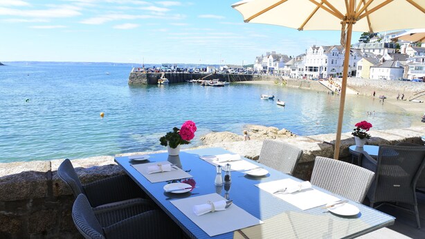 A dining table set with white tableware, a vase with pink flowers, and wicker chairs at a seaside location with a view of boats and buildings. - The Idle Rocks (Truro)