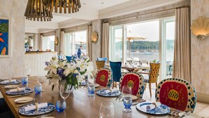 A dining area with a wooden table set for a meal, large windows with curtains, and a view of the sea. - The Idle Rocks (Truro)