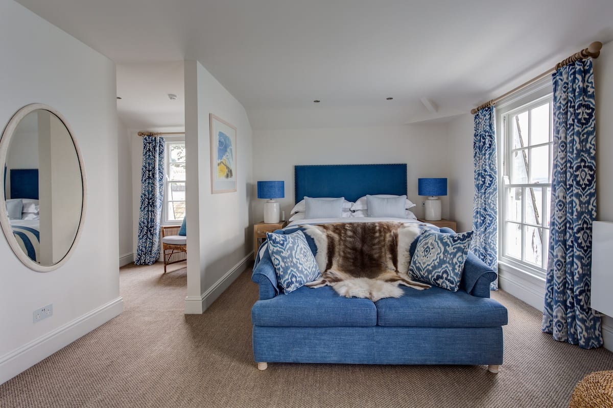A bedroom with a large bed, a blue sofa, a round mirror, and a window with patterned curtains.