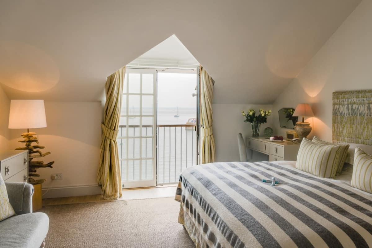 A bedroom with a bed, a desk, a chair, a lamp, and a view of the sea through a balcony door.