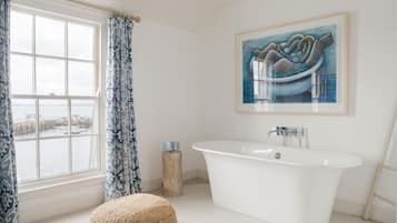 A bathroom with a freestanding bathtub, a large window with a view of the sea, and a painting above the bathtub.