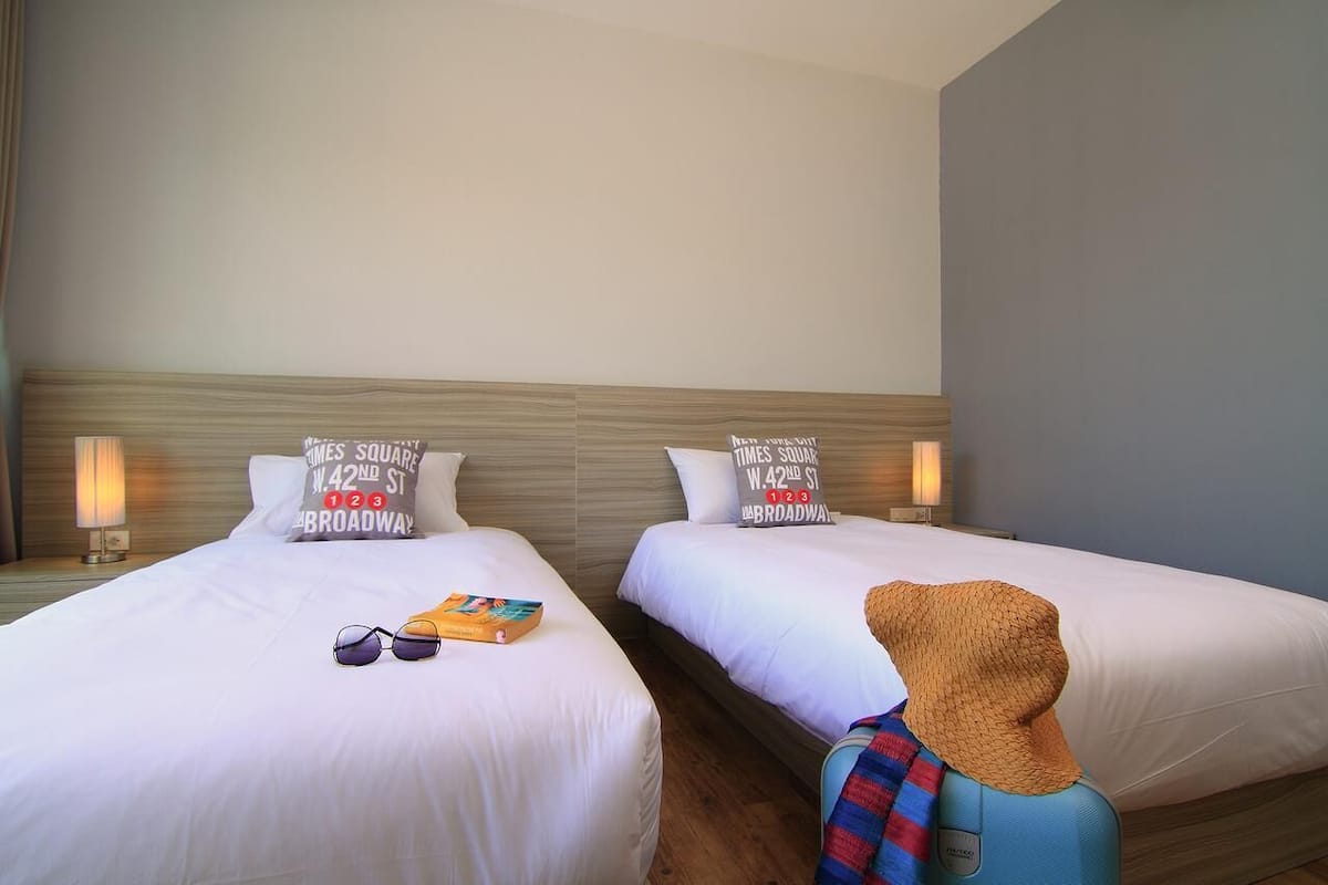 Une chambre d’hôtel avec deux lits simples, chacun orné d’un oreiller sur lequel figurent les mots « Times Square » et « Broadway ». Sur le lit de gauche, on trouve une paire de lunettes de soleil et un livre, et sur le lit de droite, un chapeau.