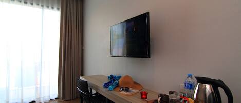 Habitación de hotel con un escritorio de madera, televisor montado en la pared, ventana con cortinas y una bandeja con diversos objetos, incluyendo una tetera y una botella de agua.