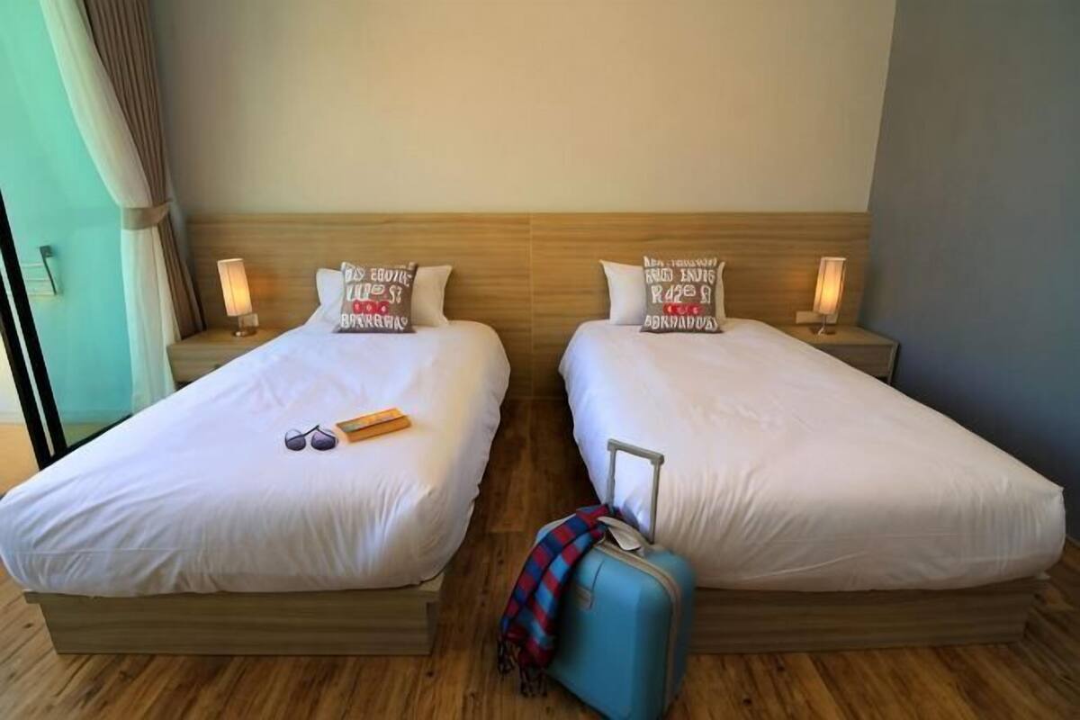 Deux lits simples avec des têtes de lit en bois, des tables de chevet avec des lampes et une valise bleue posée sur le sol.