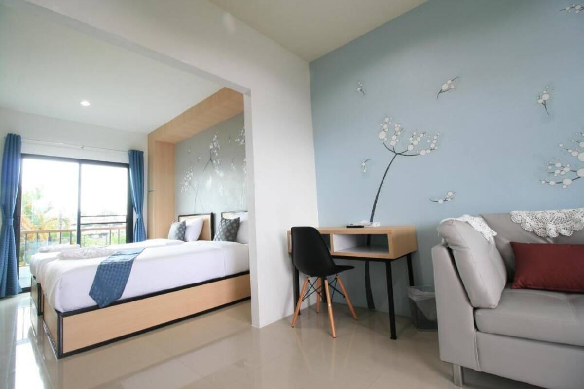 A modern hotel room with a bed, desk, and sofa, a balcony with curtains, and a wall with floral wall art.