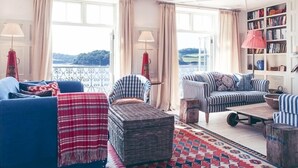 A cozy living room with a blue sofa, a striped armchair, and a patterned rug. There is a bookshelf filled with books and a view of the outdoors through large windows. - St Mawes Hotel (Truro)