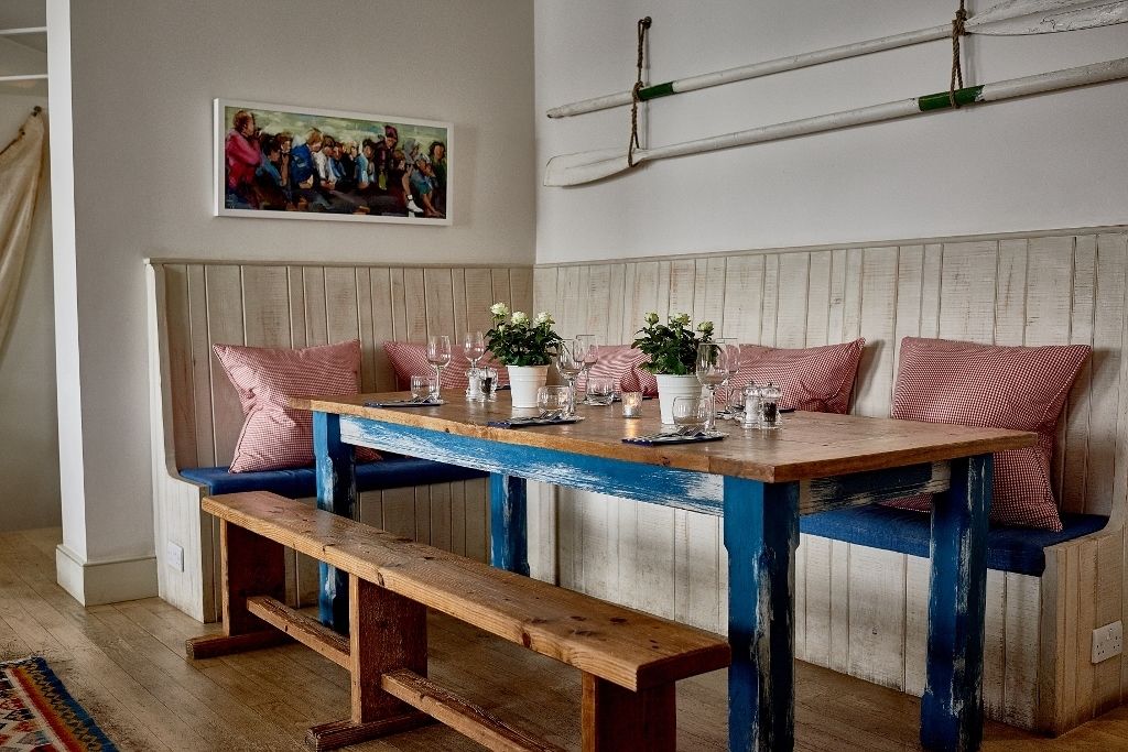 A dining area with a wooden table set for four, benches with red and white checkered cushions, and a wall-mounted surfboard.