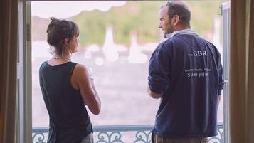Un homme et une femme se tiennent près d’une fenêtre, d’où l’on peut voir un plan d’eau et de la verdure.