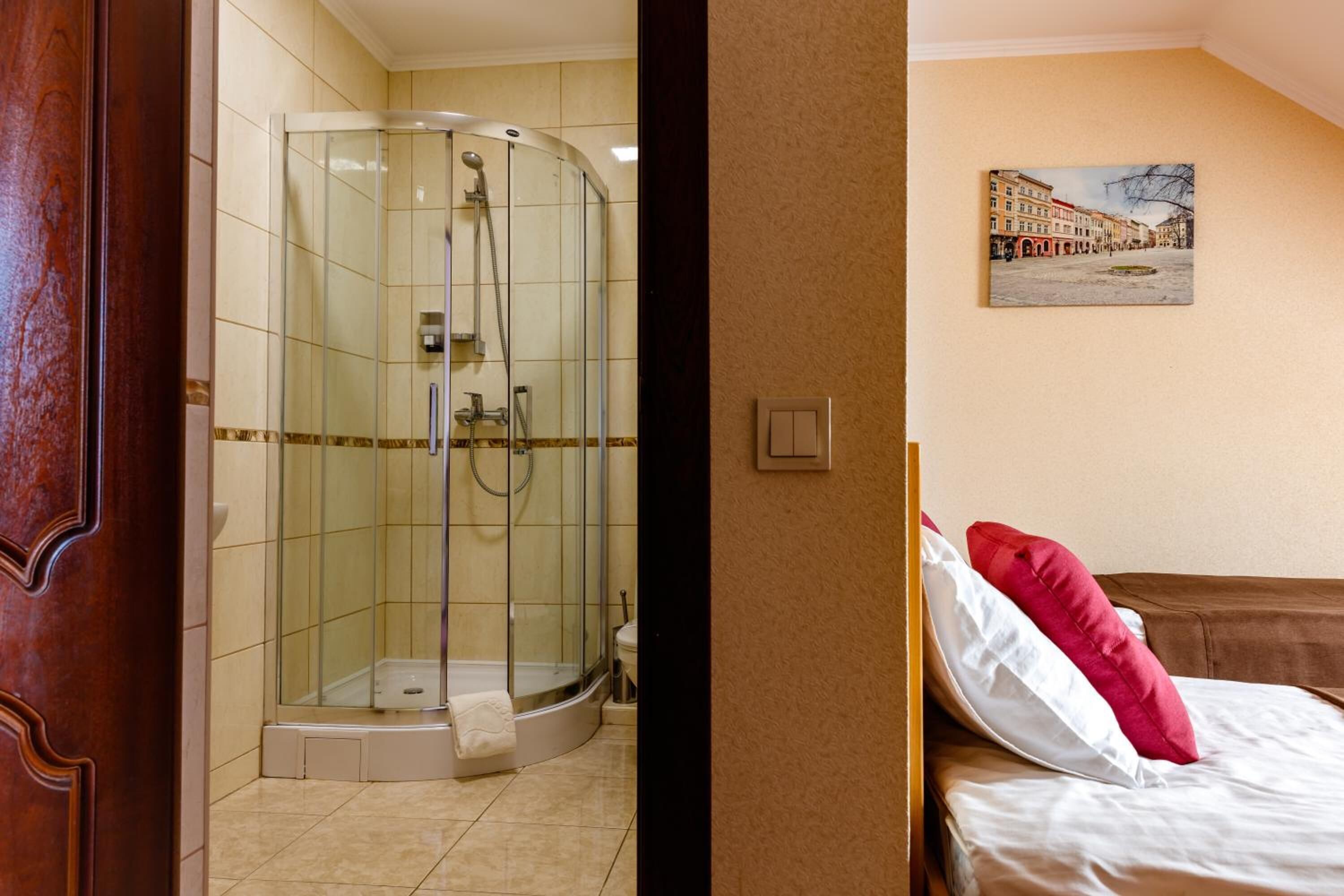 A hotel room with a glass-enclosed shower, a bed with white and red pillows, and a wall-mounted painting of a street scene.