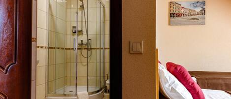 A hotel room with a glass-enclosed shower, a bed with white and red pillows, and a wall-mounted painting of a street scene.