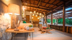 Un espace de vie chaleureux et décloisonné, avec un plafond en bois rustique, une table à manger agrémentée d’une lampe, et une vue sur une végétation luxuriante à l’extérieur.