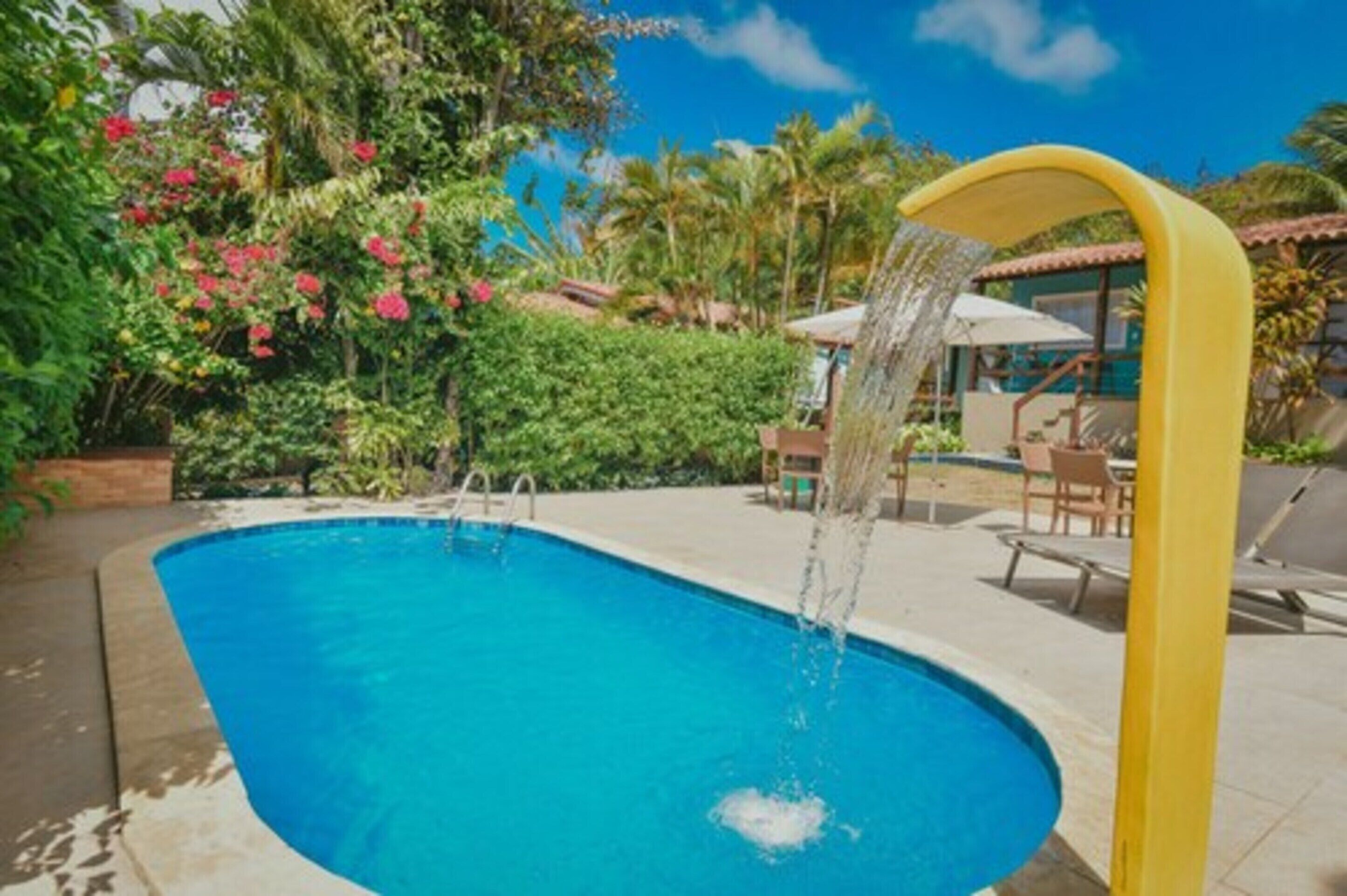 A swimming pool with a water feature, surrounded by lush greenery and a clear blue sky.