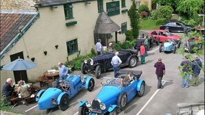A vintage car event with classic cars parked in a row, a building with a sign reading "The Ivy Inn," and people gathered around.