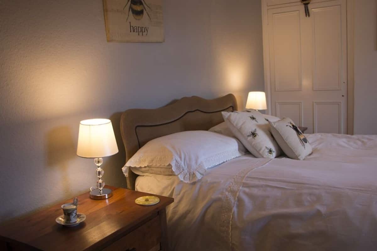 Ein ordentlich gemachtes Bett mit weißen Leinentüchern und dekorativen Kissen, ein Nachttisch mit Lampe und ein an der Wand hängendes Kunstwerk mit dem Wort 'happy'.