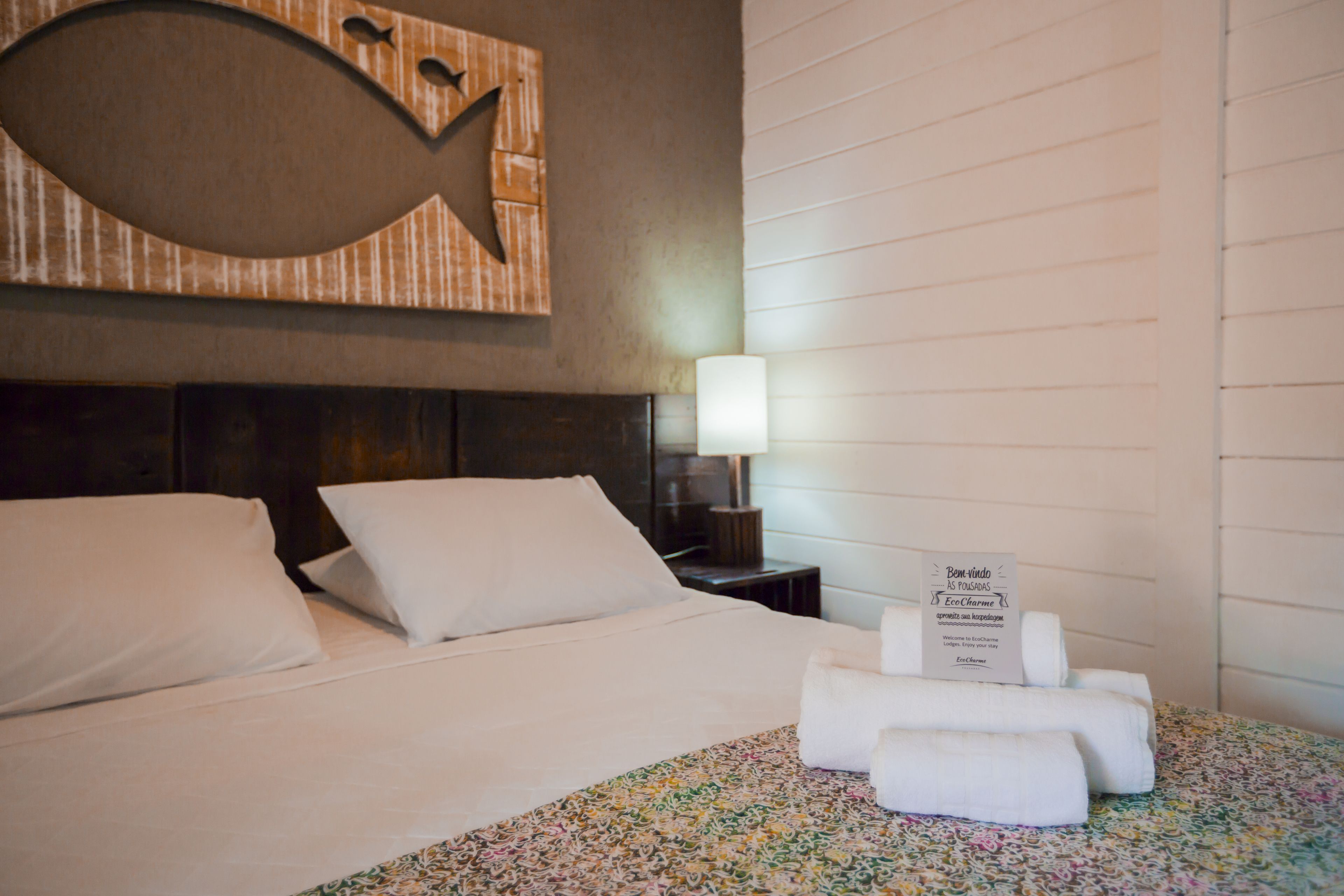 A hotel room with a neatly made bed, a wall-mounted artwork, a bedside lamp, and a welcome sign.