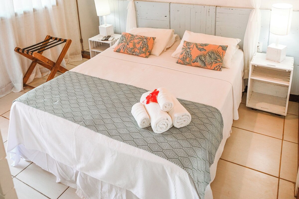 Un dormitorio con cama, cortinas blancas, techo de madera, un cuadro de un pez y una mesita de noche con lámpara.